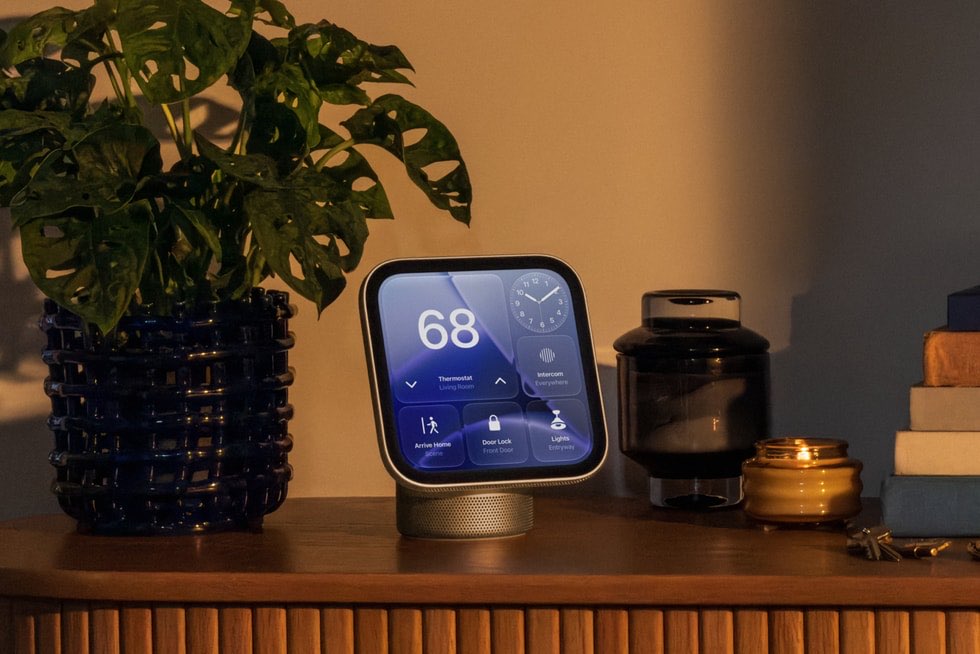 A HomeView on a wooden sideboard at dusk, beside a large plant, a lit candle and a stack of books. Its screen shows a thermostat at 68 degrees, a clock, and buttons for Intercom, a door lock and lights.