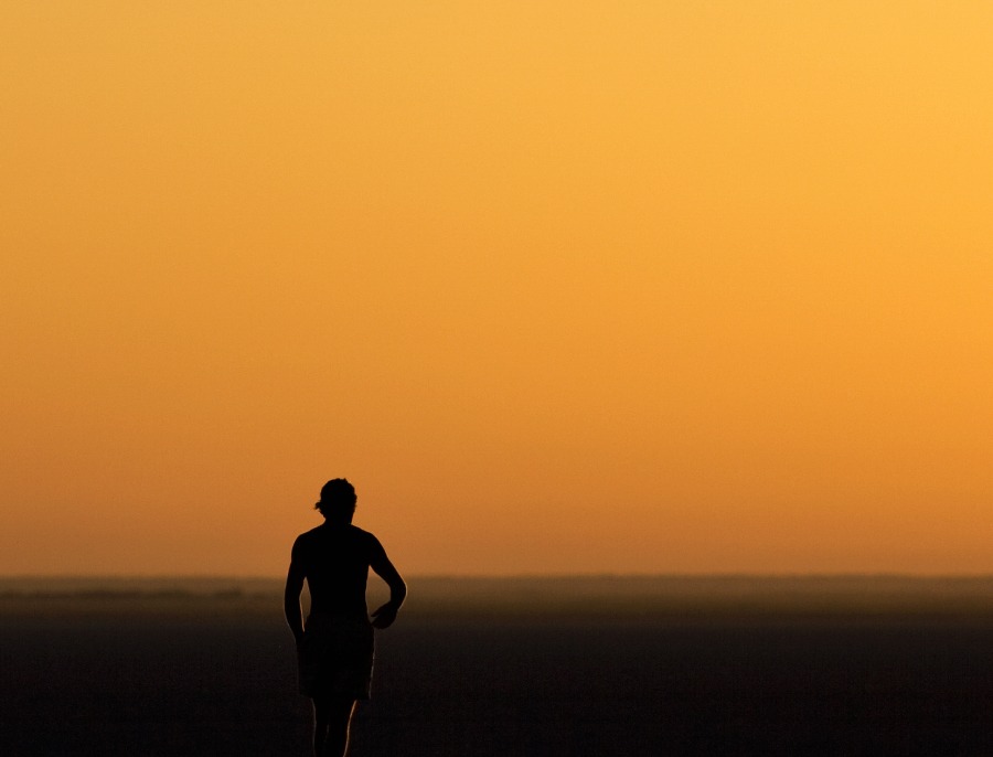 A lone figure walking across flat open ground under a wide orange sky at sunset