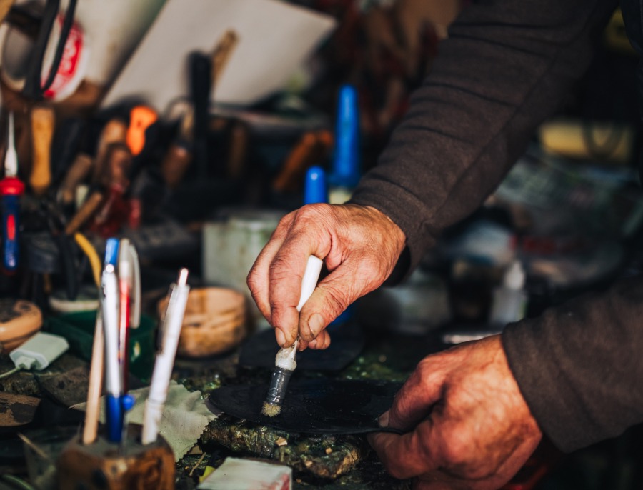 Hands working at a cluttered workbench crowded with brushes, hand tools and jars